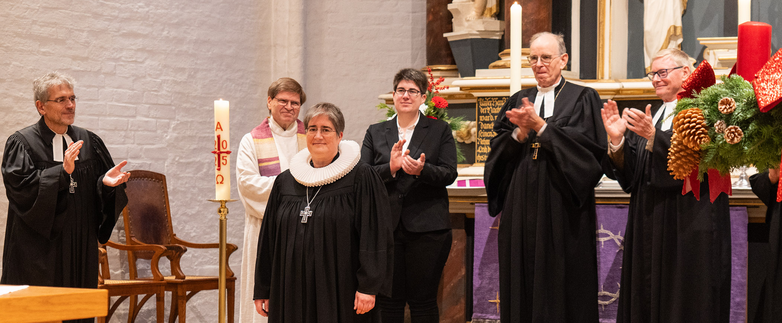 Mehrere Personen, teils in Talaren, stehen im Halbkreis hinter einer weiblich gelesenen Person im Talar.