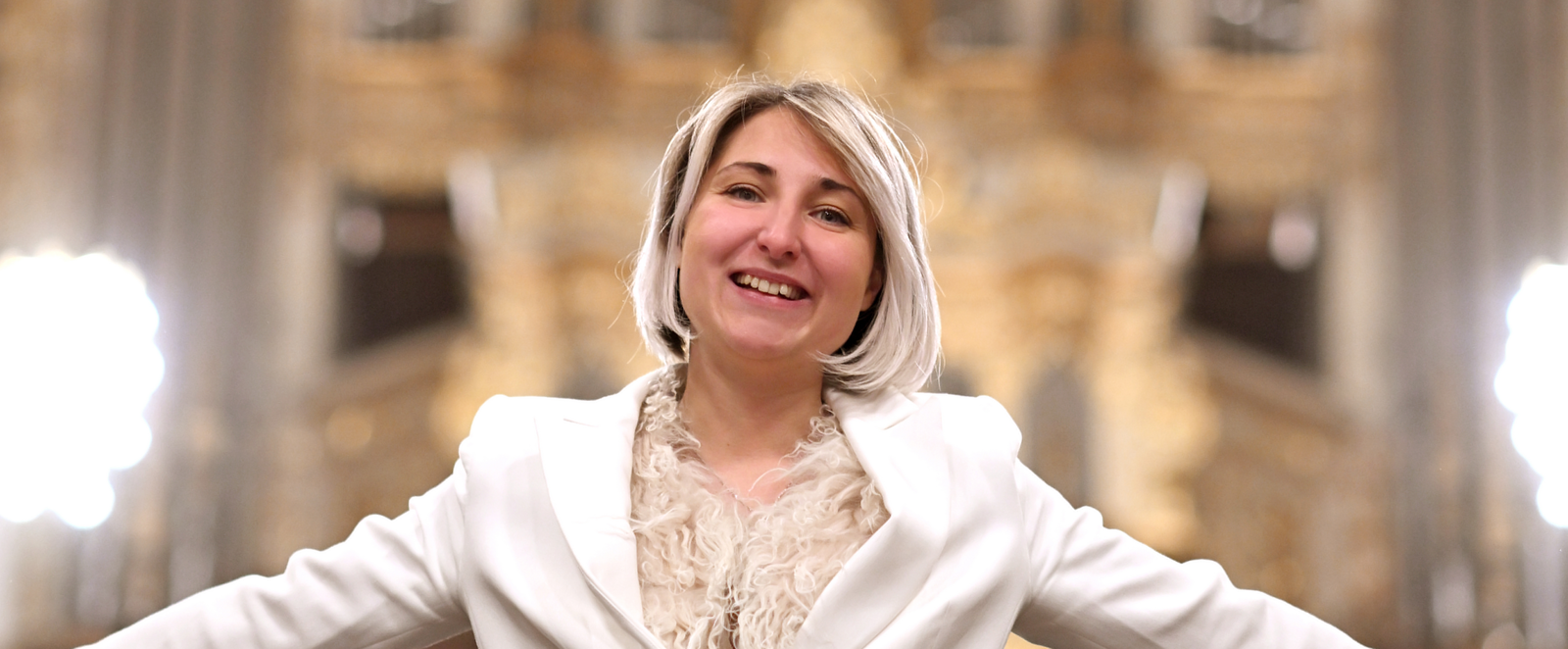 Eine weiblich gelesene Person in weißem Anzug strahlt vor einer goldenen Orgel.
