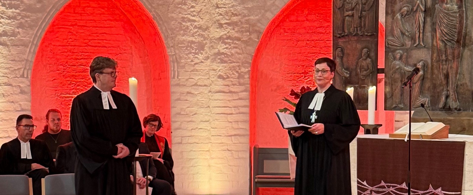 Zwei Personen in Talaren in einer rot erleuchteten Kirche.