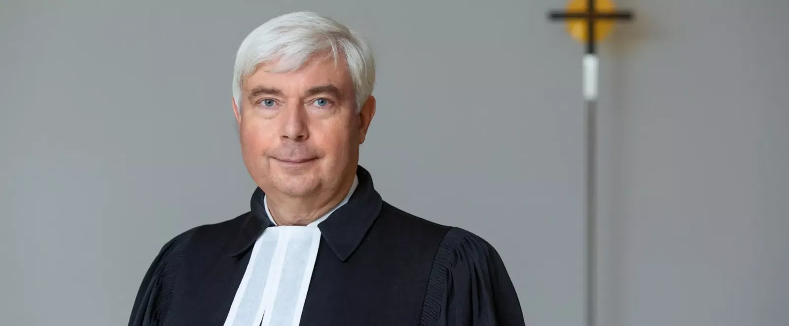 Eine als Mann lesbare Person mittleren Alters steht im Talar vor einem Kreuz.