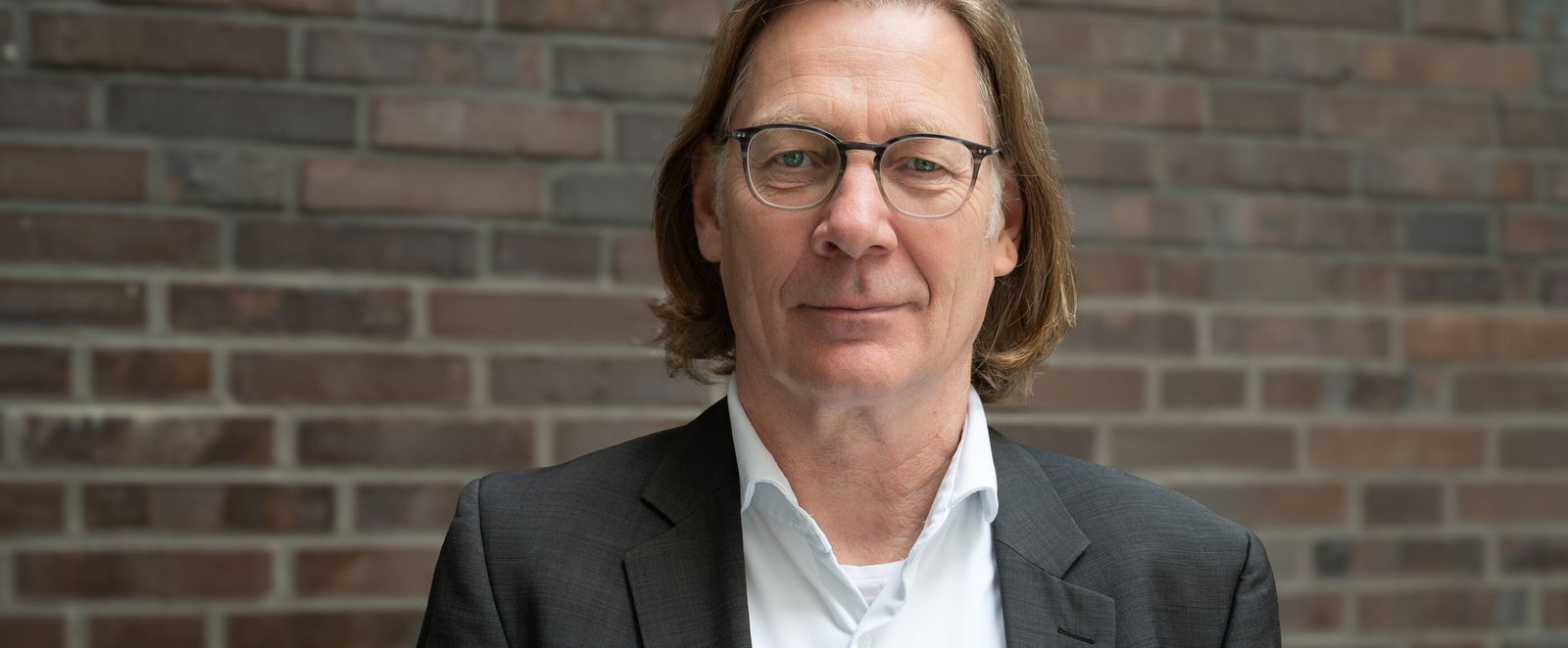 Eine männlich gelesene Person mit mittellangen Haaren, Brille, weißem Hemd und dunklem Sakko.