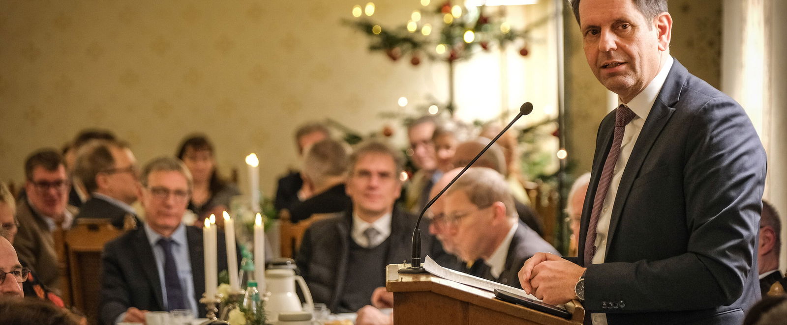 Eine männlich zu lesende Person steht an einem Redepult und spricht zu Menschen, die an einem festlich gedeckten Tisch sitzen.