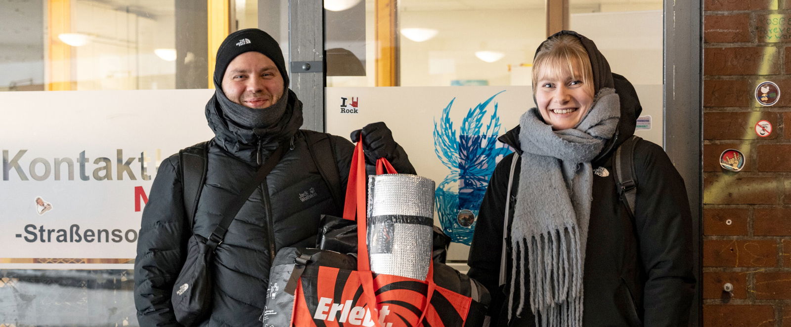 Eine als Mann und eine als Frau lesbare Person in Winterkleidung mit einem Beutel mit Schlafsack und Isomatte.