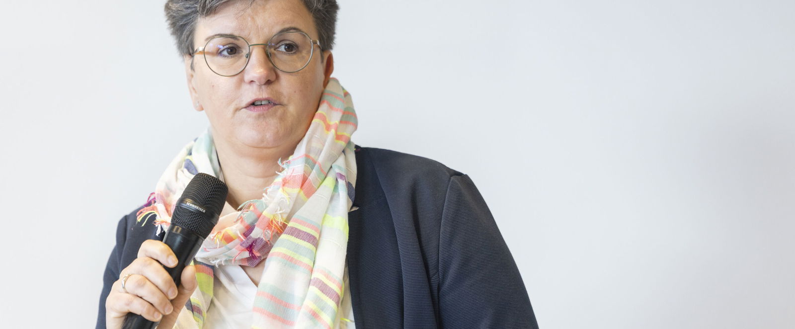 Eine weiblich gelesene Person mit blauer Jacke und Schal hält ein Mikro in der Hand.