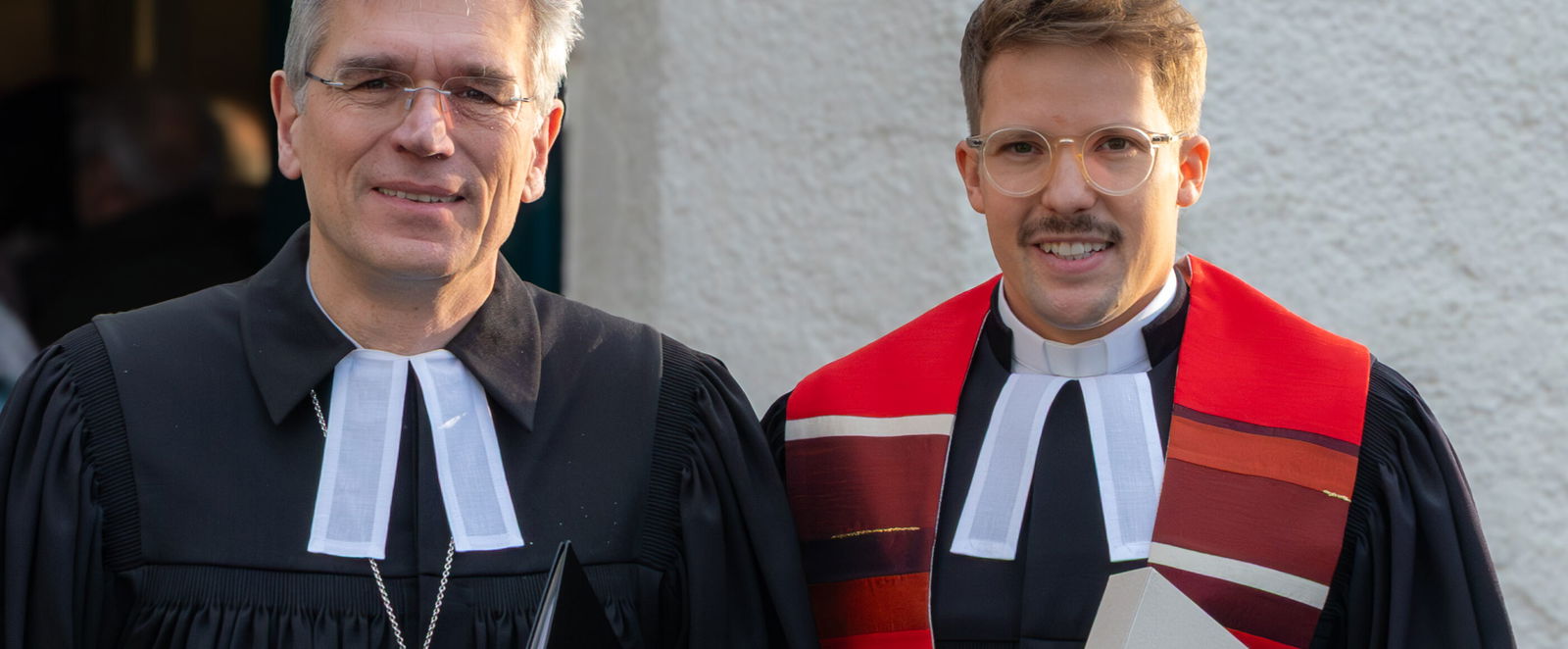 Zwei männlich gelesene Personen mit Talaren vor einer Kirche.