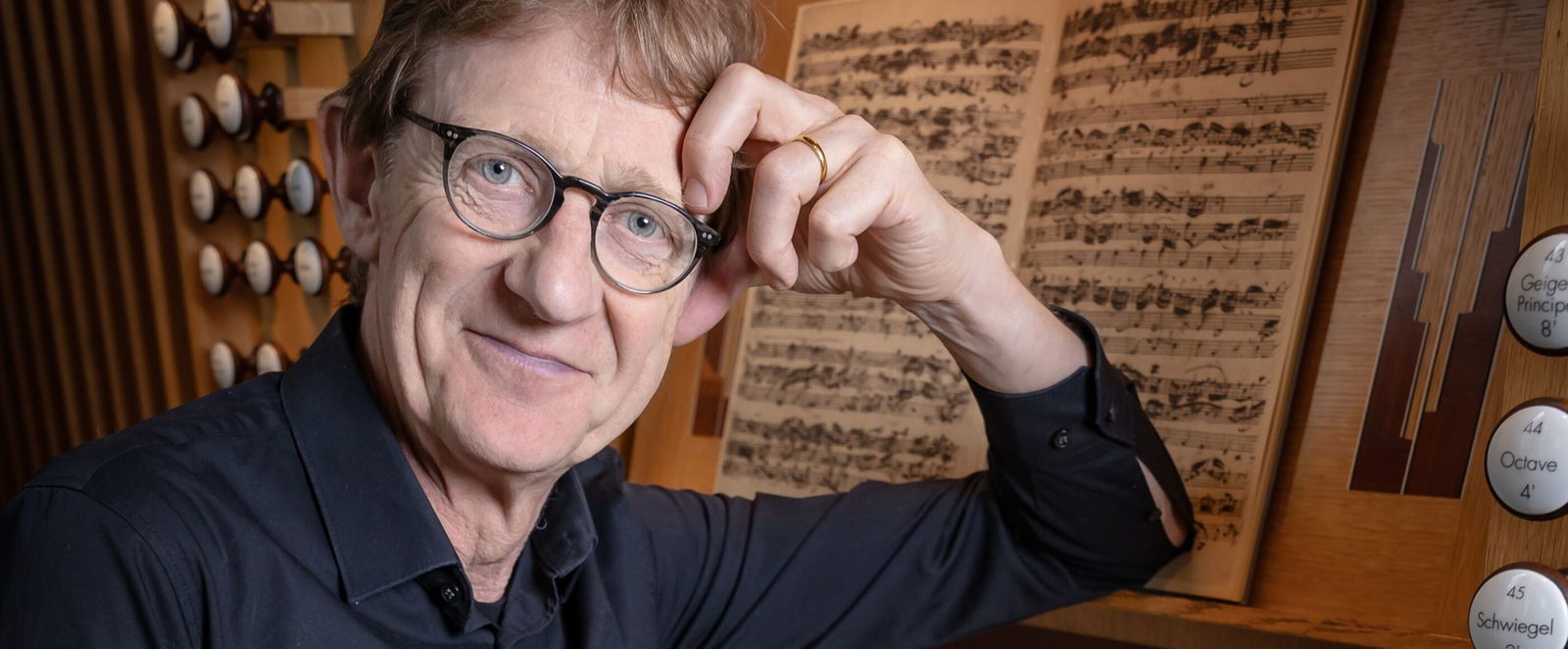 Eine männlich gelesene Person in blauem Hemd stützt sich mit einem Arm an einer Orgel ab.