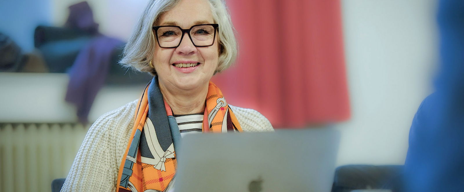 Eine weiblich gelesene Person mit mittellangen, grauen Haaren und buntem Halstuch sitzt an einem Laptop.