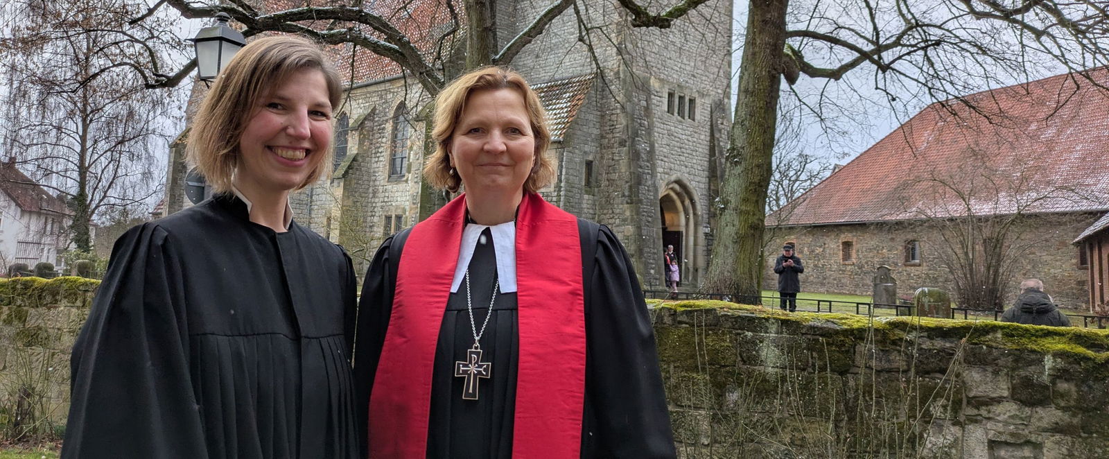 Zwei Frauen im Talar stehen vor einer Kirchenmauer.
