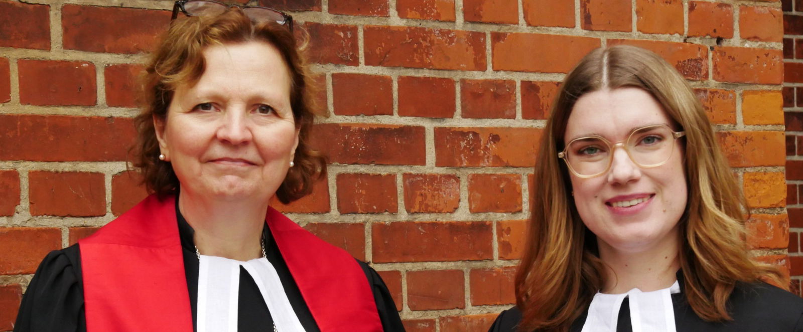 Zwei Frauen stehen im Talar vor einer Mauer.