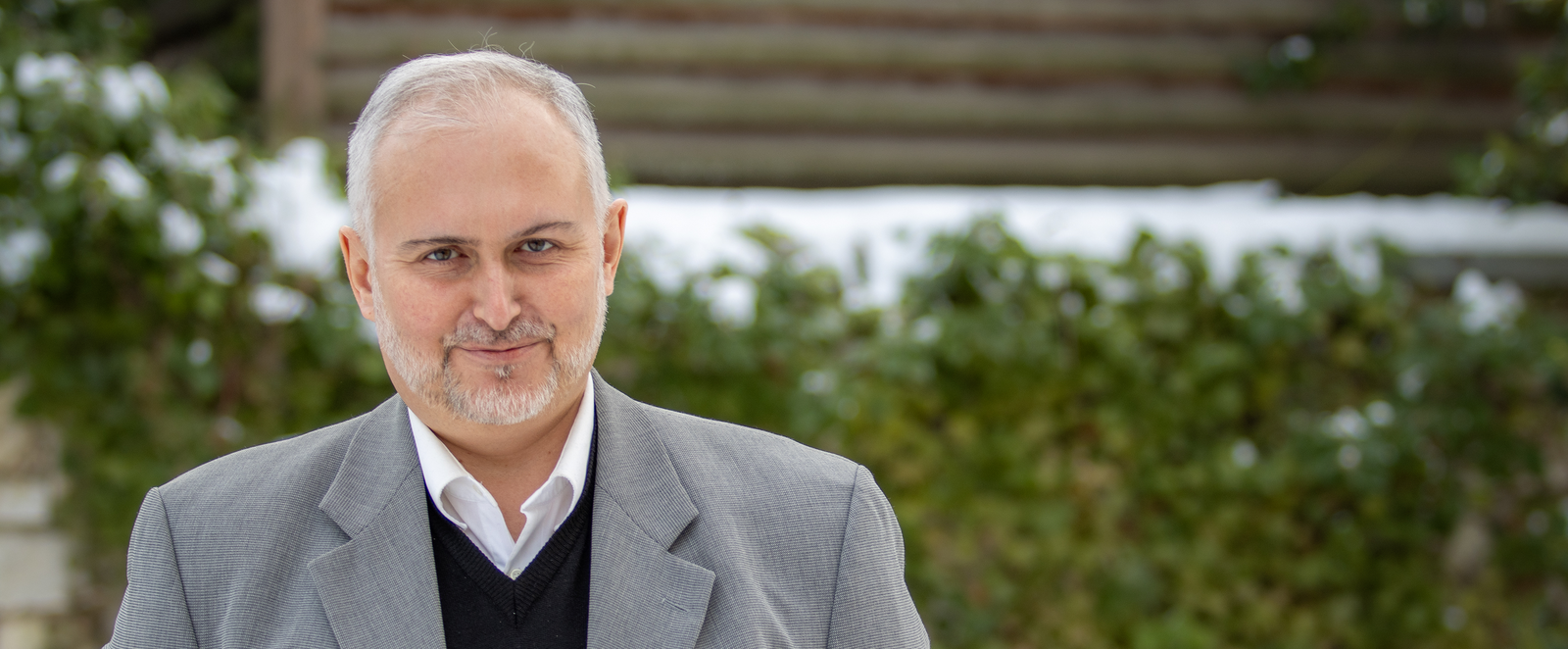 Eine männlich gelesene Person in grauem Jackett vor einer Hecke.