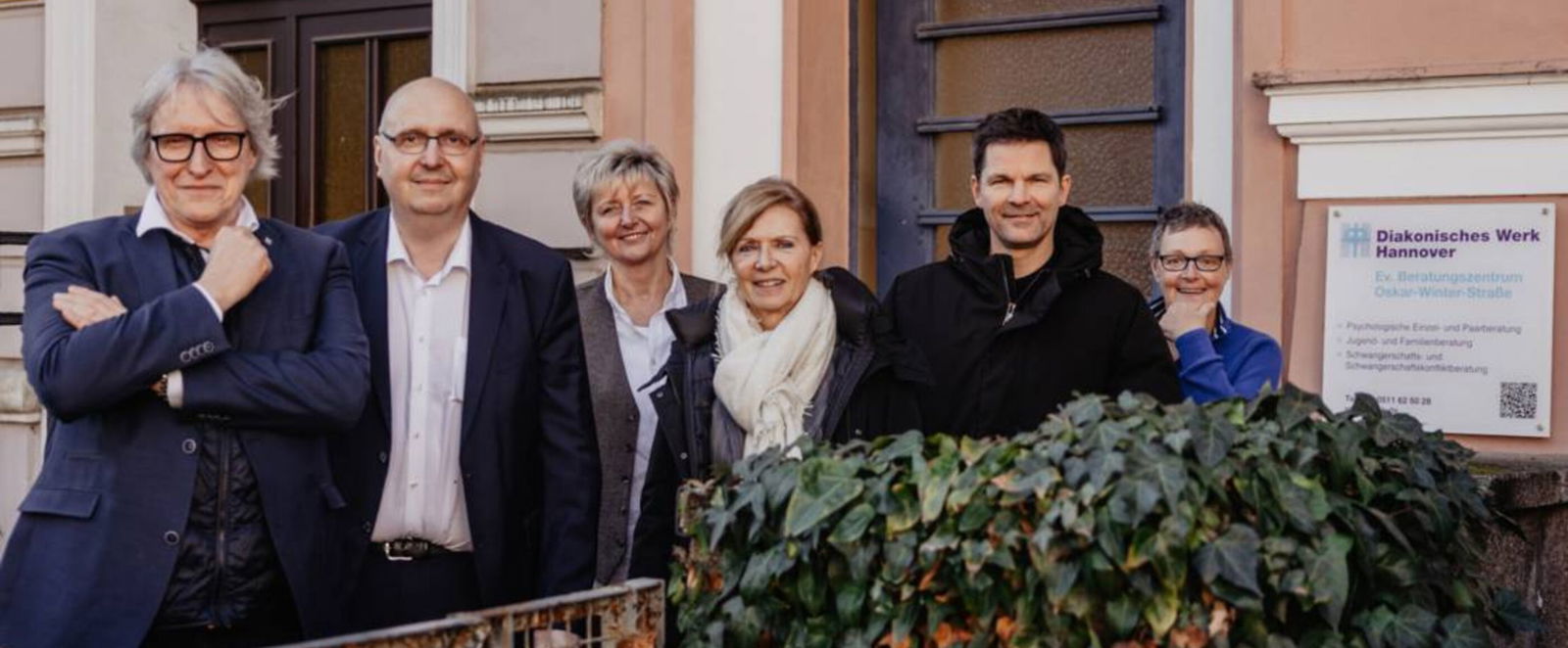 Eine Gruppe aus als Männer und Frauen lesbare Personen steht vor einem Haus der Diakonie.