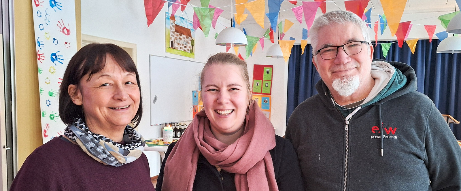 Drei Personen stehen lächelnd zusammen in einem bunt geschmückten Raum.