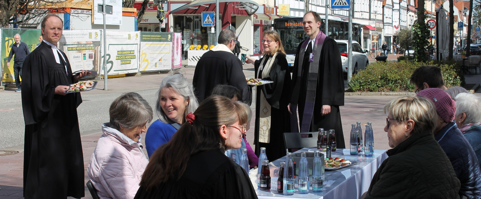 Mehrere Personen sitzen an einem gedeckten Tisch im Freien, einige Personen, teils in Talaren stehen darum.
