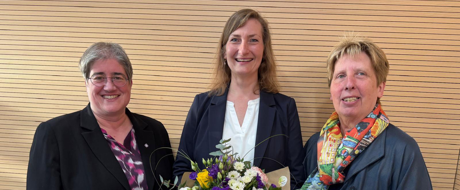 Drei als Frauen lesbare Personen, die mittlere hält einen Blumenstrauß.