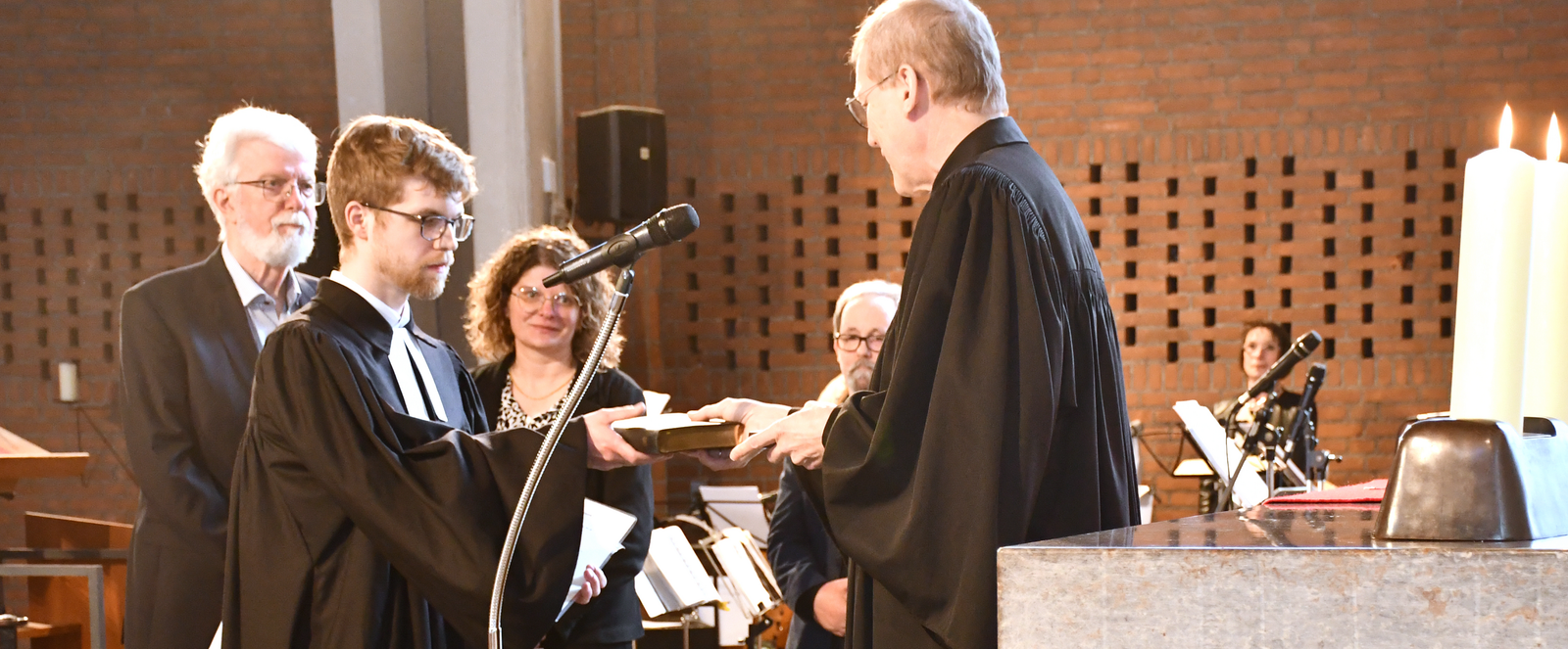 Mehrere Personen im Altarraum einer Kirche. Zwei männlich gelesene Personen in Talaren stehen sich gegenüber und überreichen eine Bibel.