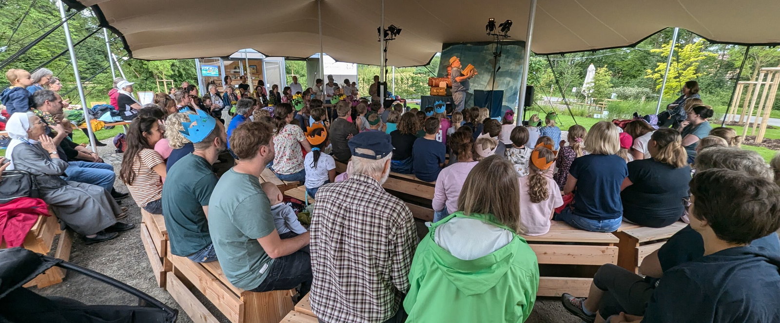 Viele Menschen sitzen unter einem Zeltdach und schauen auf eine Bühne.