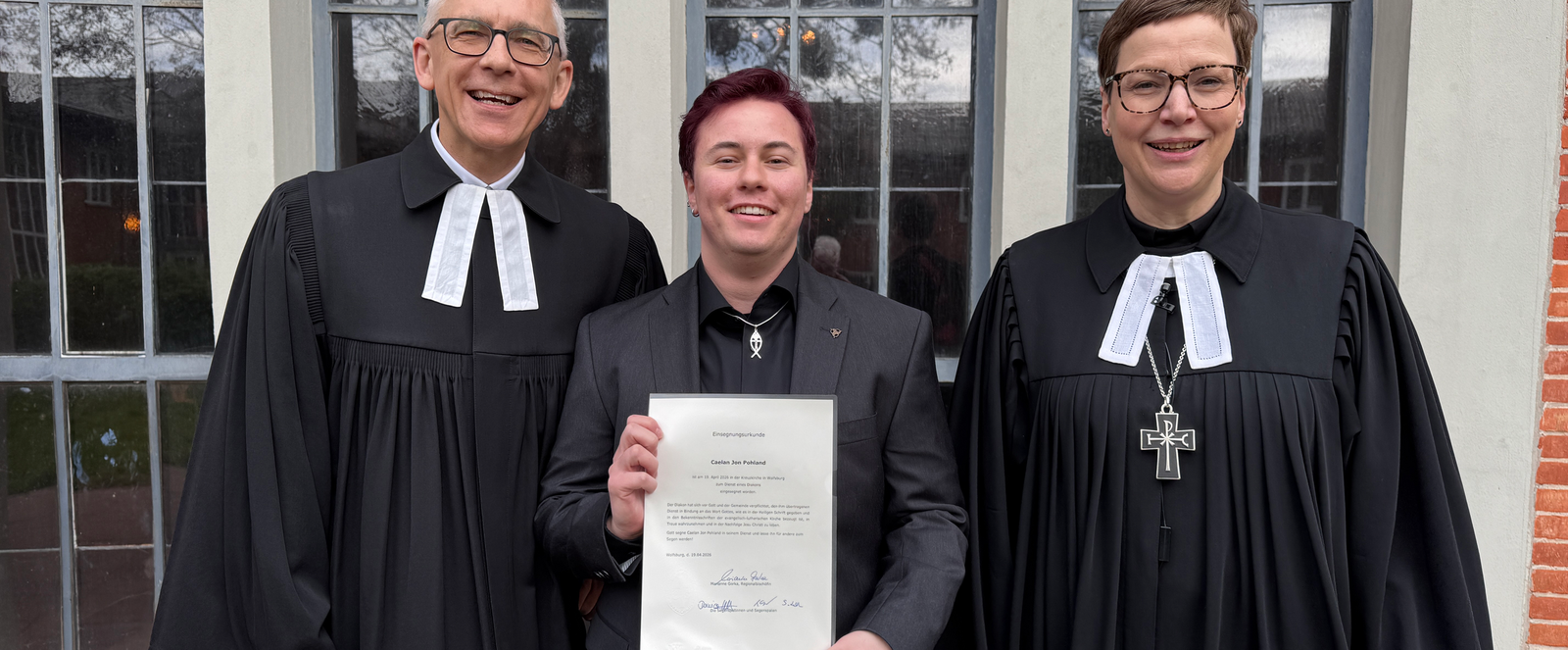 Drei Personen, zwei in Talaren, stehen zusammen, die dritte hält eine Urkunde in Händen.