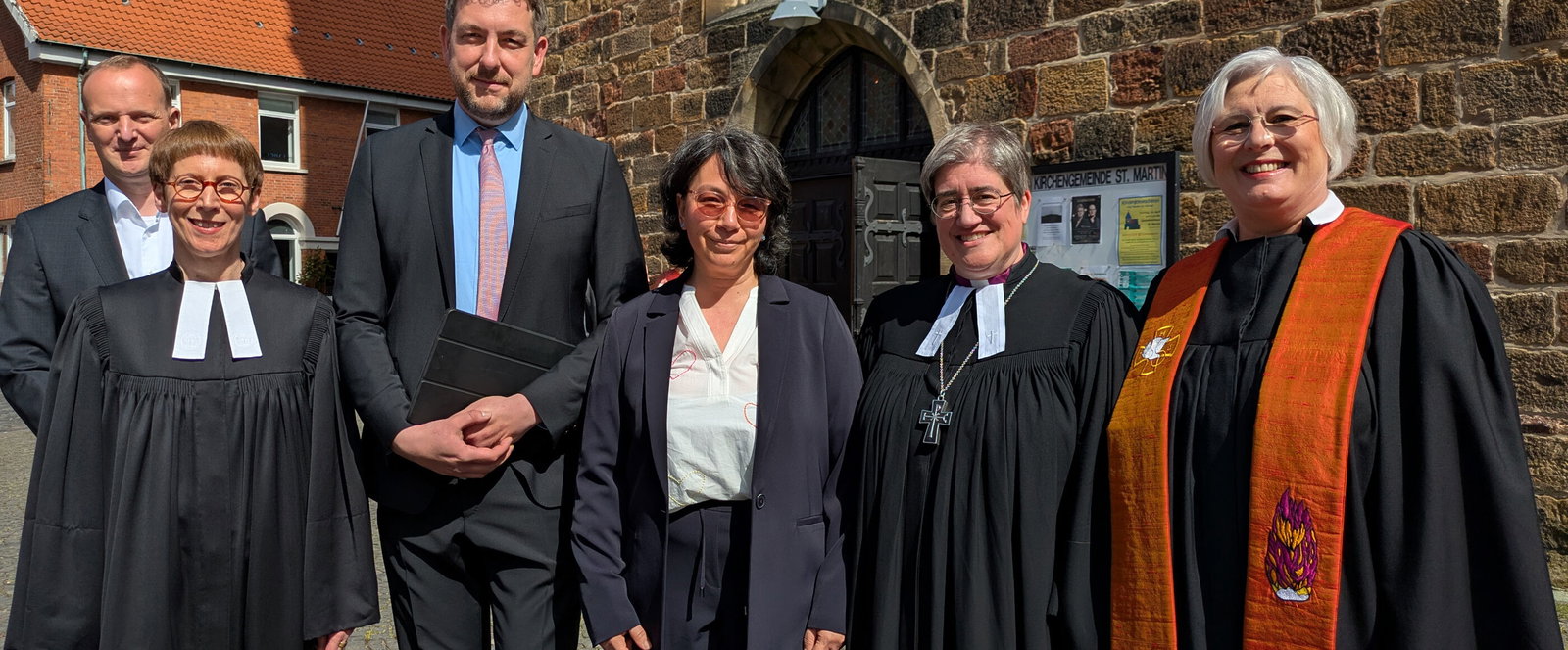 Eine Gruppe aus als Frauen und Männer lesbaren Personen, teils im Talar, vor einem Backsteingebäude.