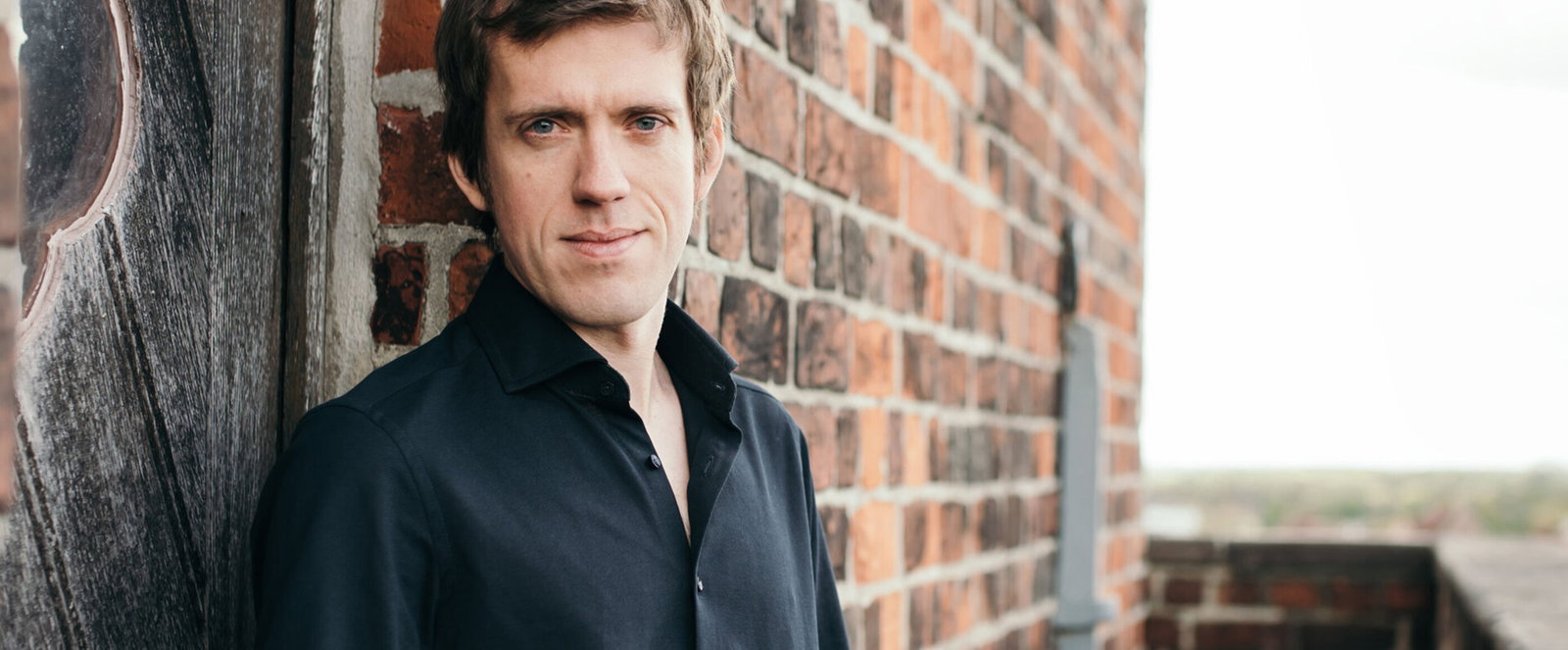 Eine als männlich lesbare Person im schwarzen Hemd lehnt an einer Backstein-Hauswand.