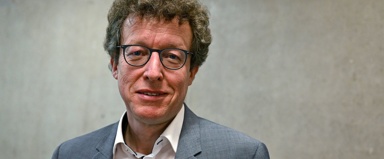 Eine als Mann lesbare Person mit Brille und Locken, steht im weißen Hemd und Sakko vor einer Betonwand.