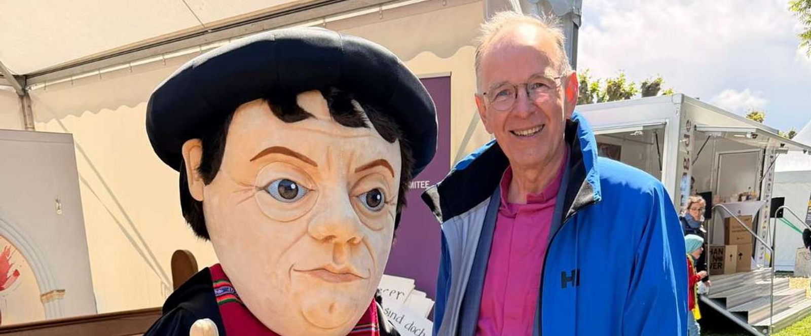 Eine männlich gelesene Person in blauer Jacke steht vor einem Zelt im Freien neben einer Figur, die Martin Luther ähnlich sieht. 