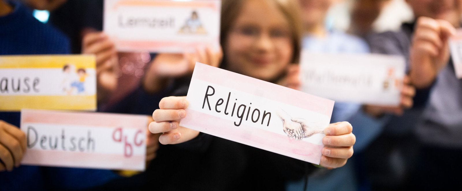 Kinder halten Schilder mit Schulfächern in die Kamera. Im Mittelpunkt steht Religion.