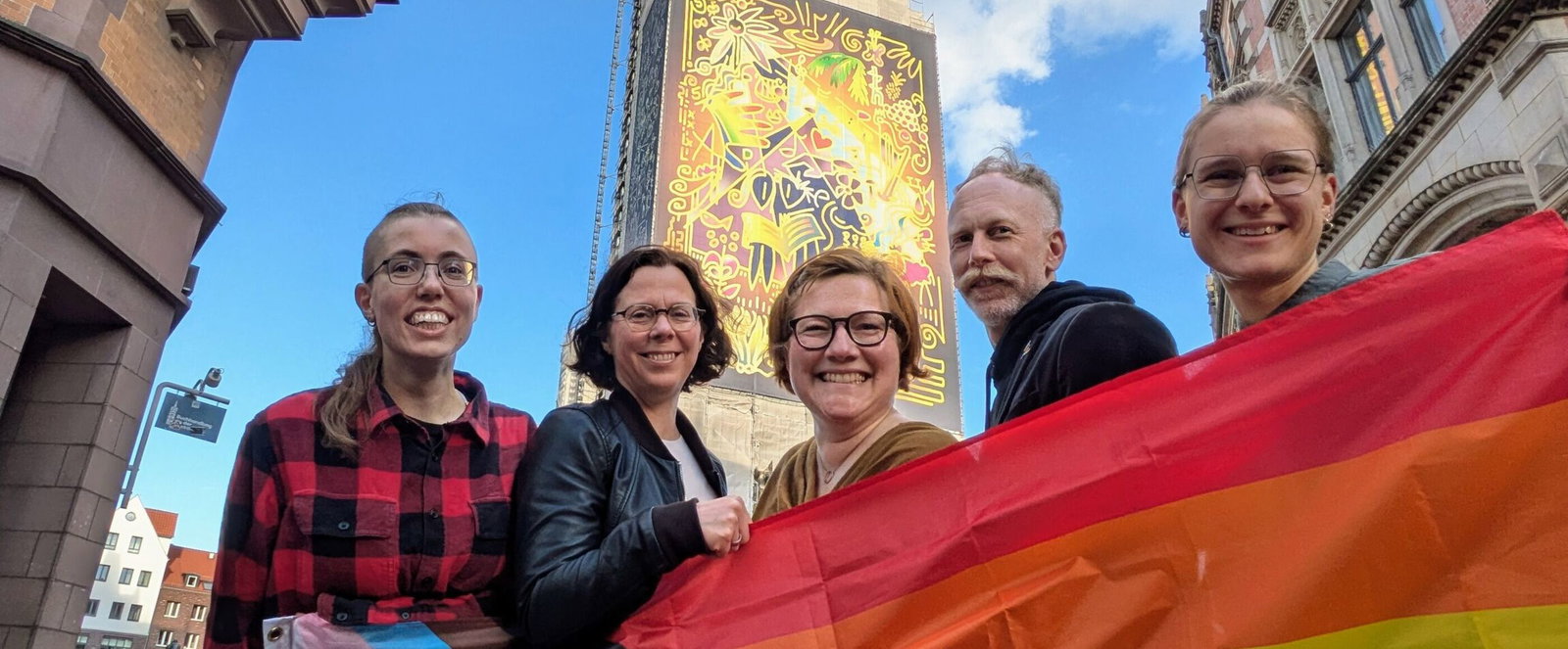 Mehrere als Männer und Frauen lesbare Personen mit einer Regenbogenflagge vor einem Kirchturm.