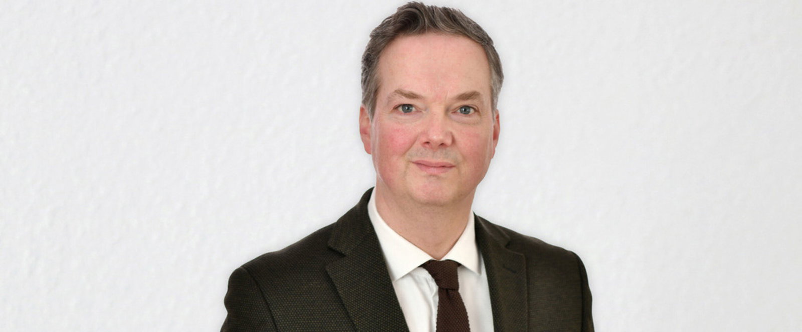 Eine als Mann lesbare Person mittleren Alters in Anzug und Krawatte vor einer weißen Wand.