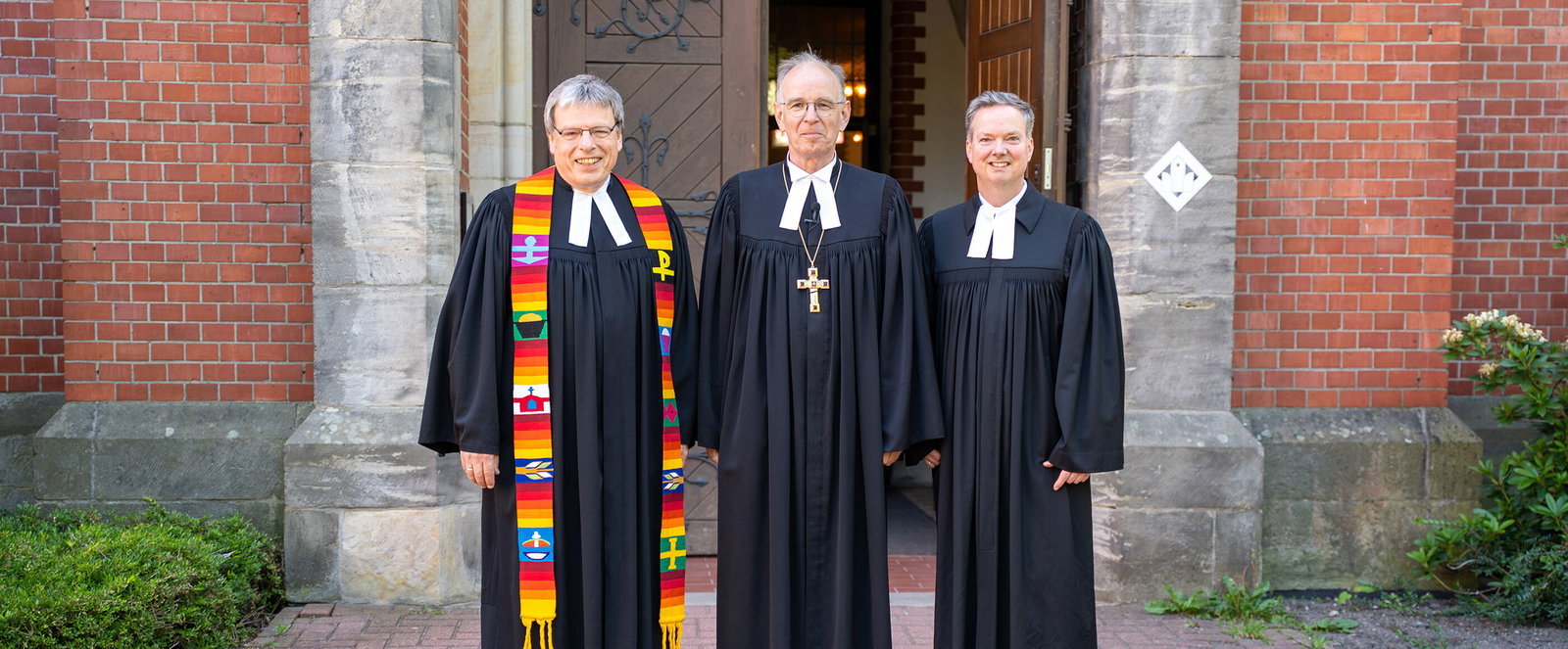 Drei als Männer lesbare Personen im Talar stehen vor der Einganstür einer Kirche.