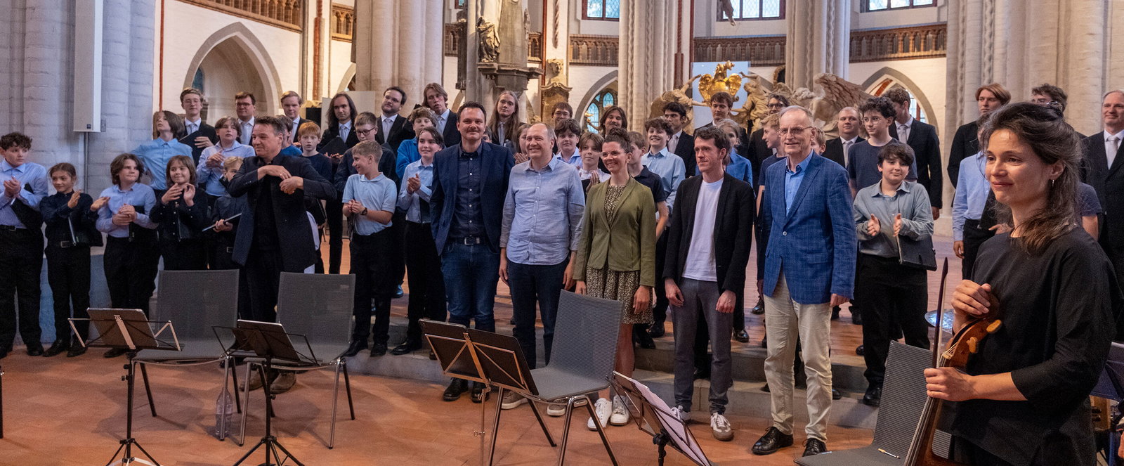 Mehrere als Frauen und Männer lesbare Personen stehen vor einem Orchester in einer Kirche.