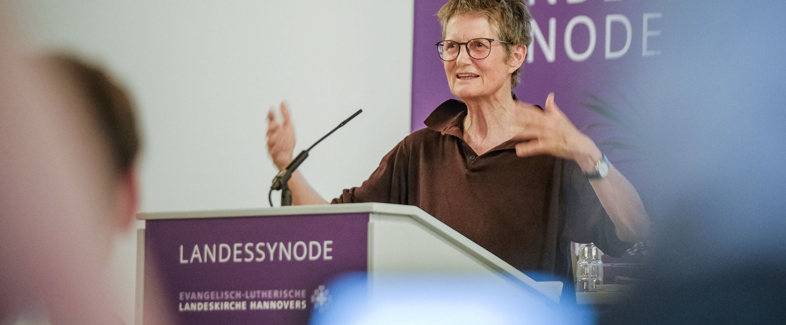 Eine als Frau lesbare Person steht an einem Redepuit und spricht gestenreich zu Publikum.