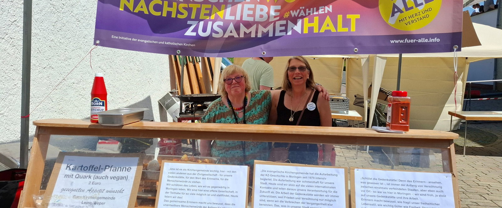 Zwei als Frauen lesbare Personen an einem Essenstand. Vorne in der Auslage liegt ein Statement zur Demokratie.