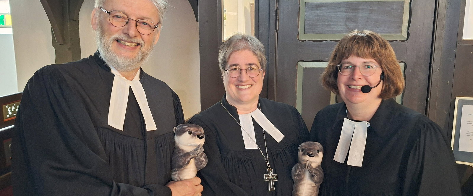 Zwei als Frauen und eine als Mann lesbare Personen im Talar halten einen Plüsch-Otter.