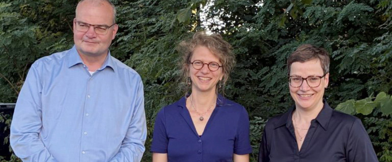 Zwei als Frauen lesbare Personen und eine als Mann lesbare Person stehen für ein Gruppenfoto vor Bäumen.