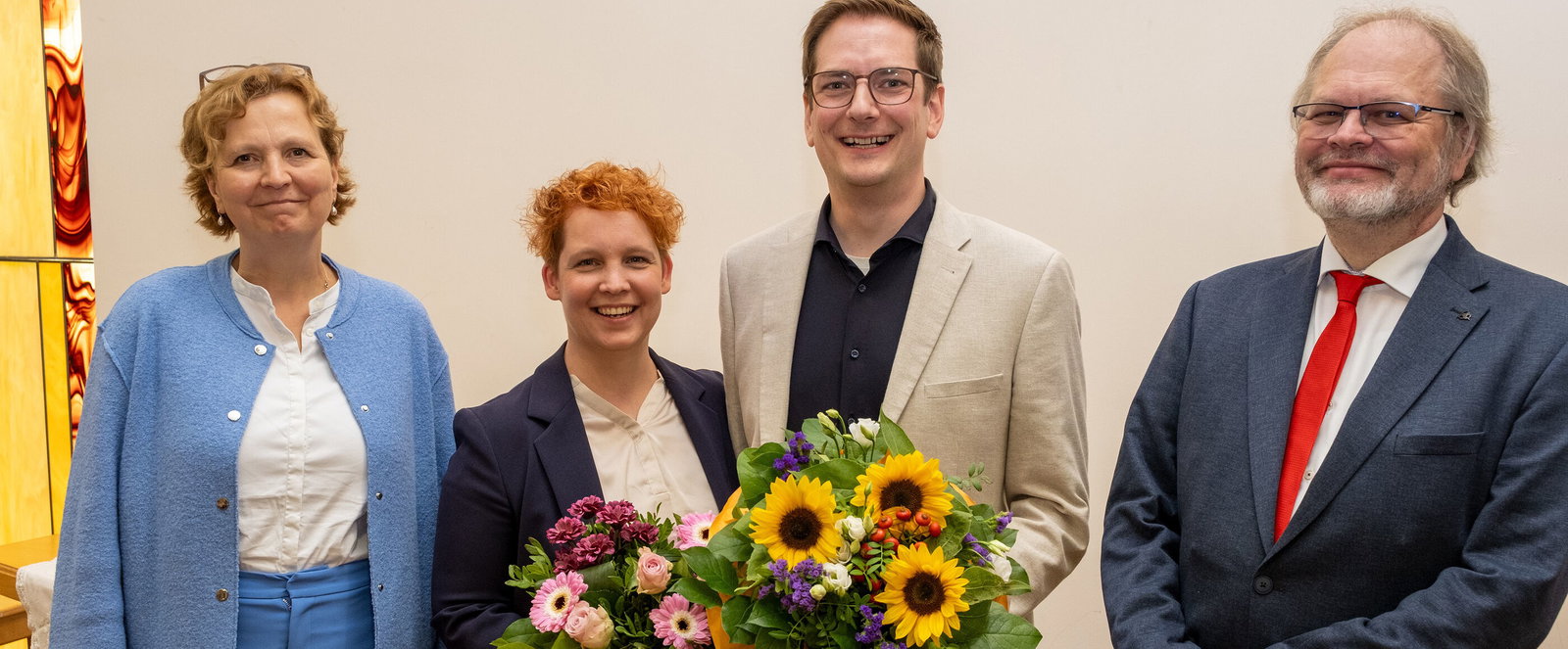Vier Menschen, zwei als Männer, zwei als Frauen lesbar, stehen für ein Gruppenfoto. Die mittleren beiden halten Blumensträuße.