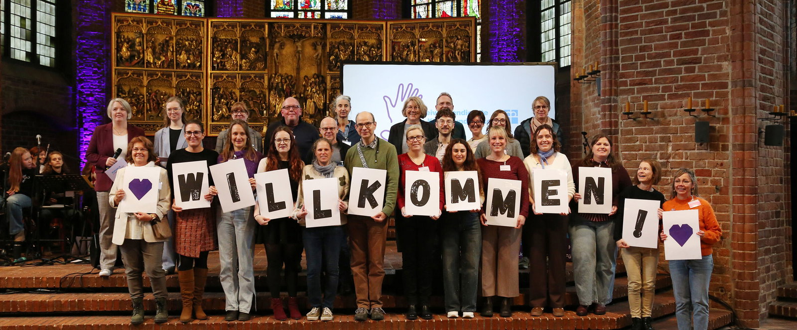 Eine große Gruppe hält in einer Kirche Buchstaben hoch, die das Wort Willkommen ergeben