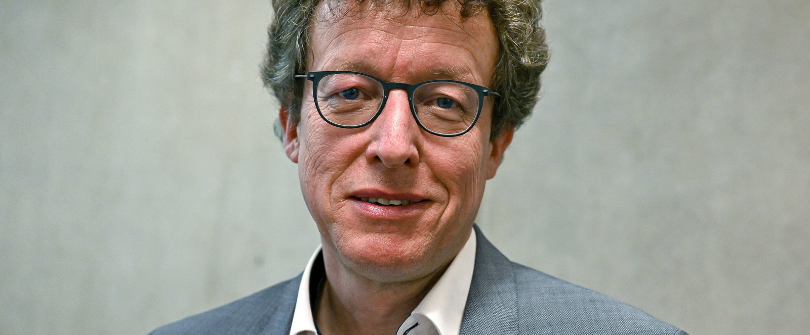 Eine als Mann lesbare Person mit Brille und Locken vor einer grauen Wand