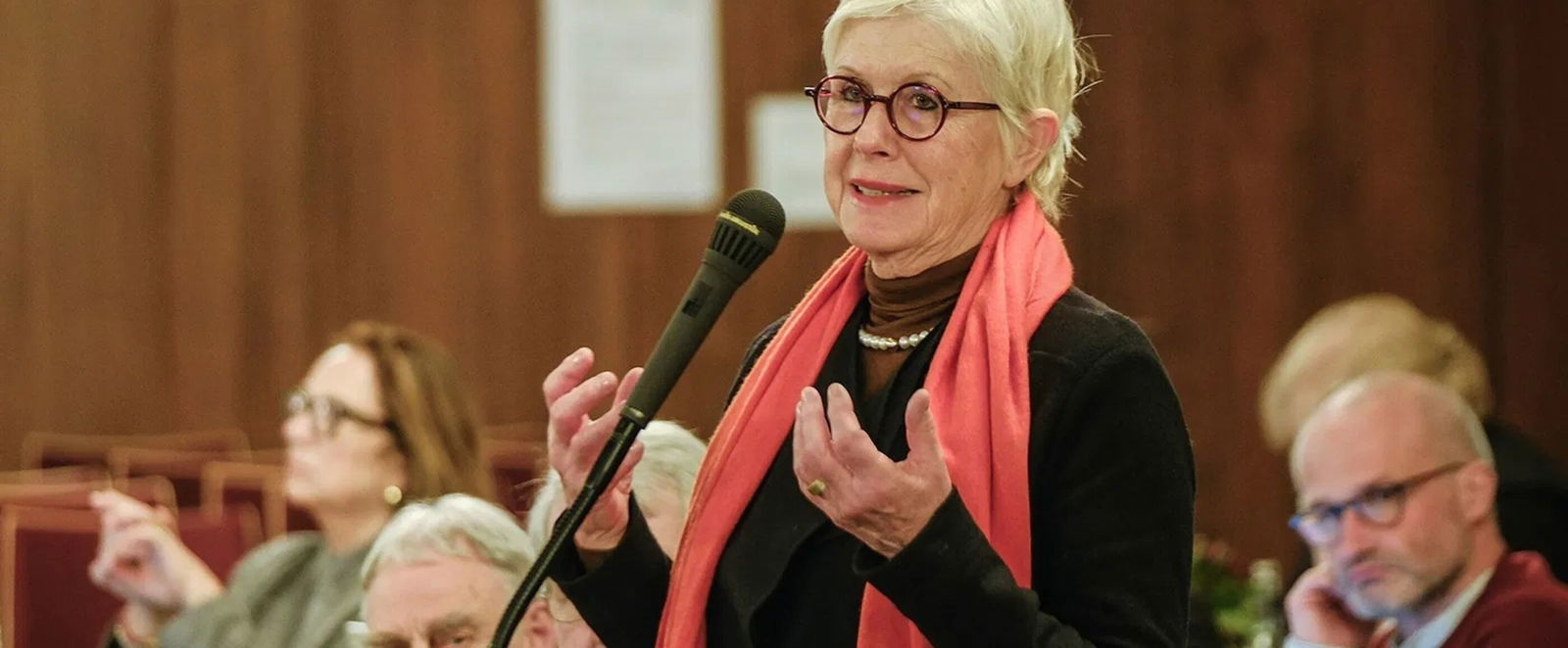 Eine als Frau lesbare Person mit rotem Schal spricht in ein Mikrofon.