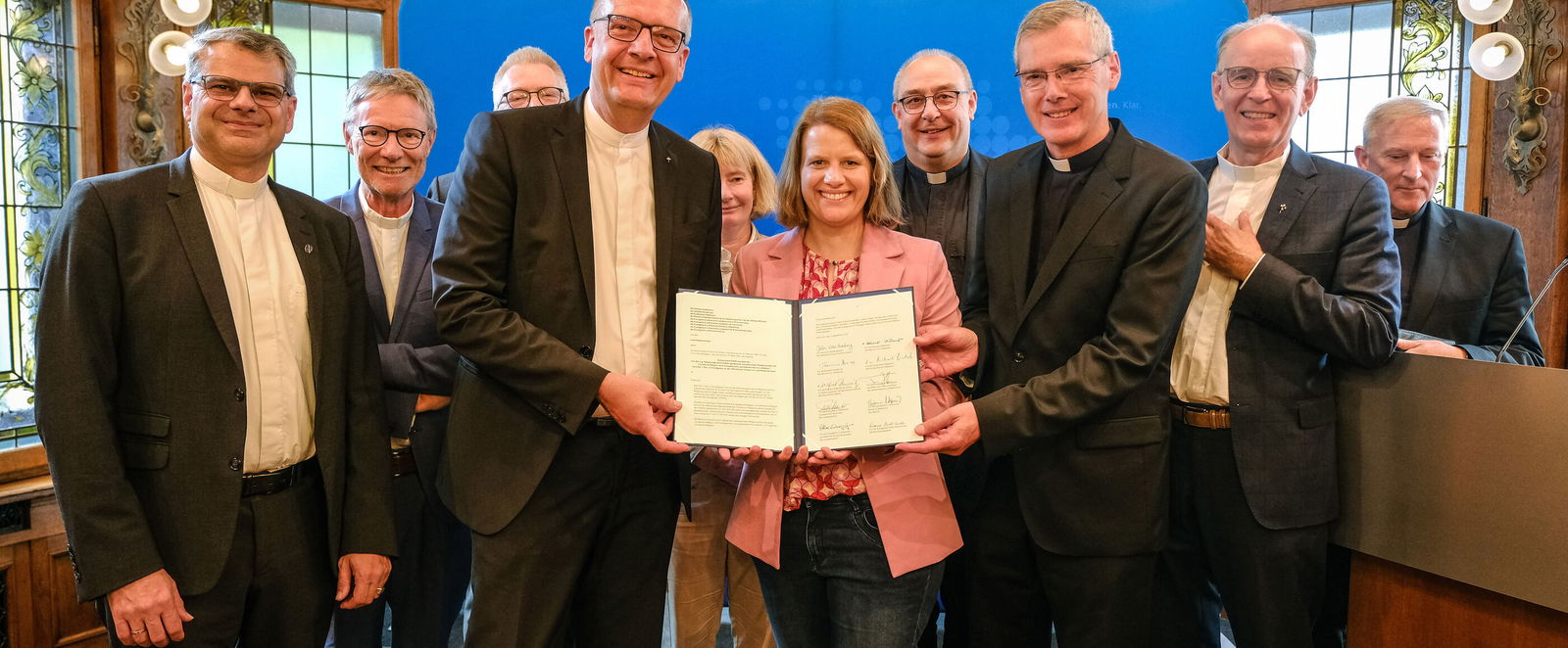 Ein Gruppe von weiblich und männlich zu lesenden Personen steht in einem Halbkreis und in der Mitte halten drei Personen ein Schriftstück in die Höhe.