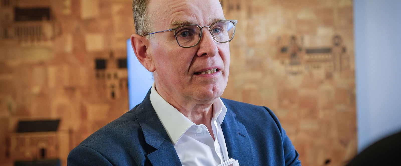 Eine männlich lesbare Person mit Brille und kurzen grauen Haaren, ohne Bart, mit blauem Sakko und weißem Hemd im Gespräch.
