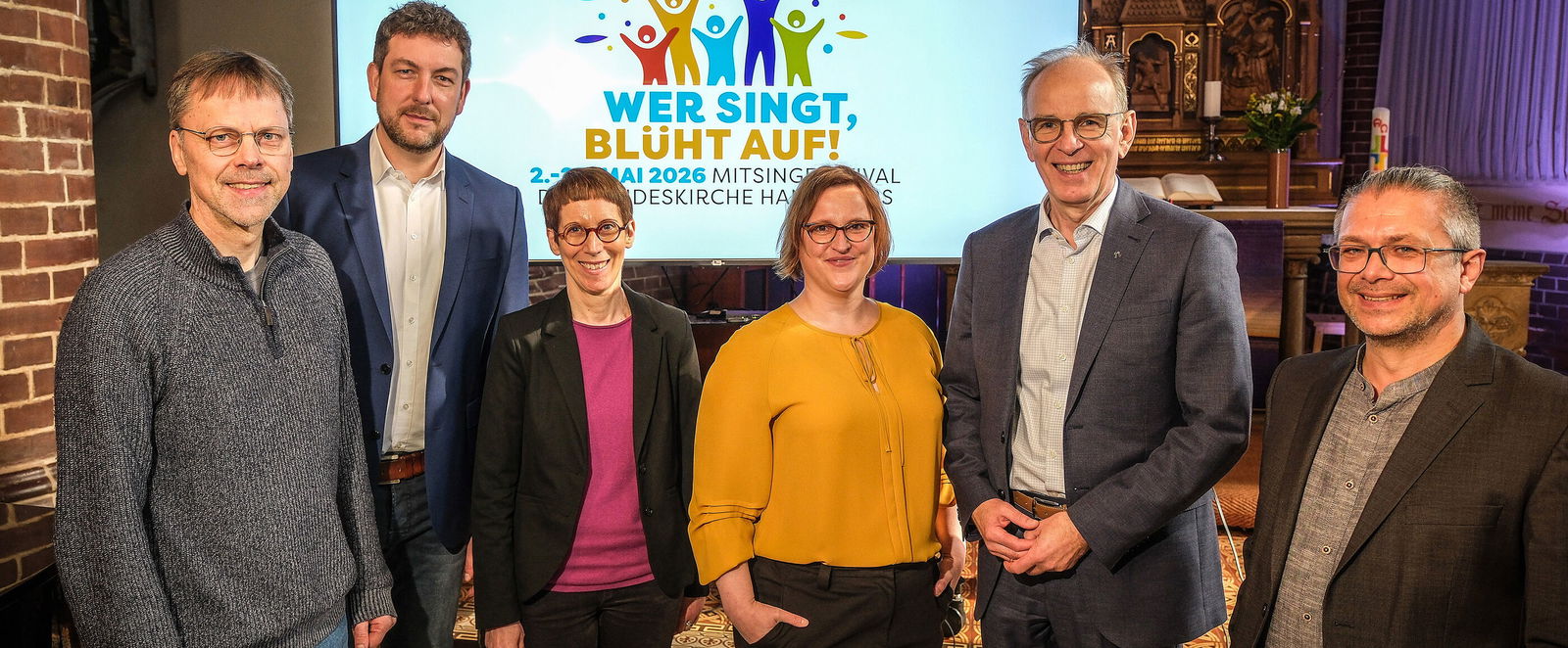 Eine Gruppe von als Männer und Frauen lesbaren Personen steht in einer Kirche vor einer Leinwand mit dem Logo zum Mitsingfestival „Wer singt, blüht auf!“