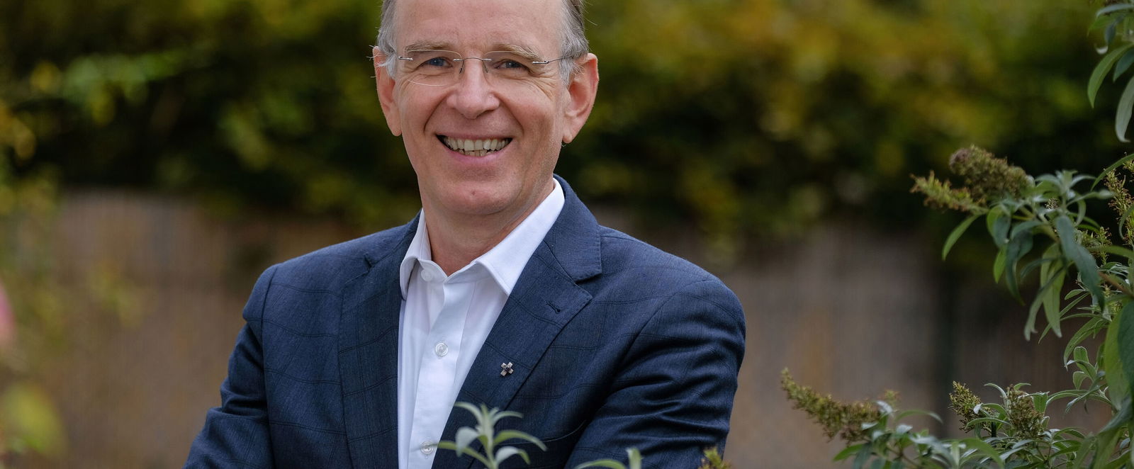 Das Bild zeigt eine männlich lesbare Person im Garten. Zu sehen ist nur der Oberkörper; die Person steht hinter einem Strauch, trägt ein dunkles Sakko und ein helles Hemd, hat kurze helle Haare und eine Brille. 