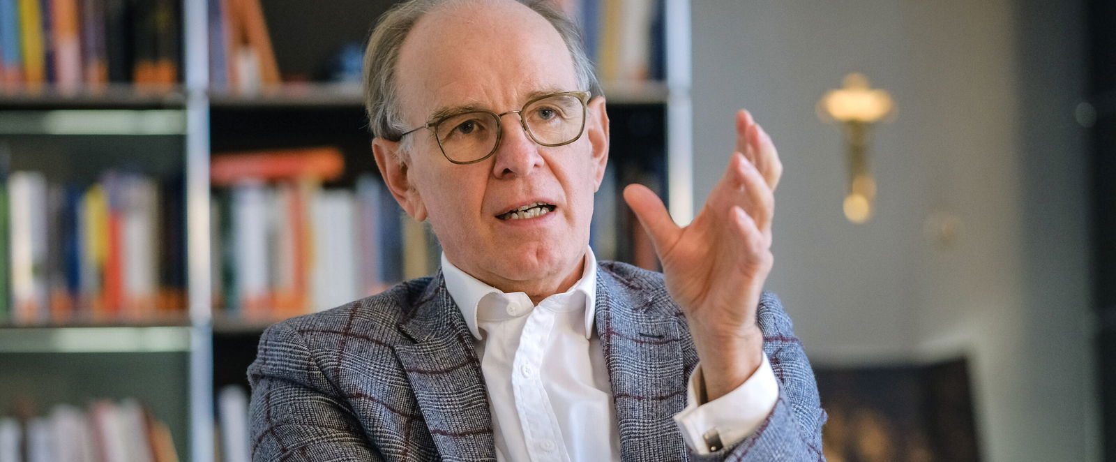 Eine männlich gelesene Person mit brauner Brillenfassung, kurzen grauen Haaren, grau-kariertem Jackett und hellblauem Hemd sitzt im Gespräch vor einem Bücherregal.