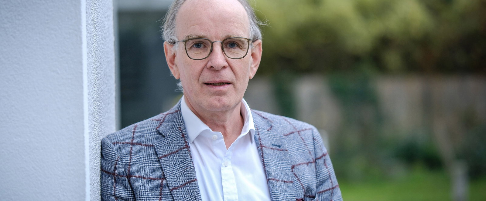 Eine männlich gelesene Person mit brauner Brillenfassung, kurzen grauen Haaren, grau-kariertem Jackett und hellblauem Hemd steht draußen und lehnt an einem Pfosten.