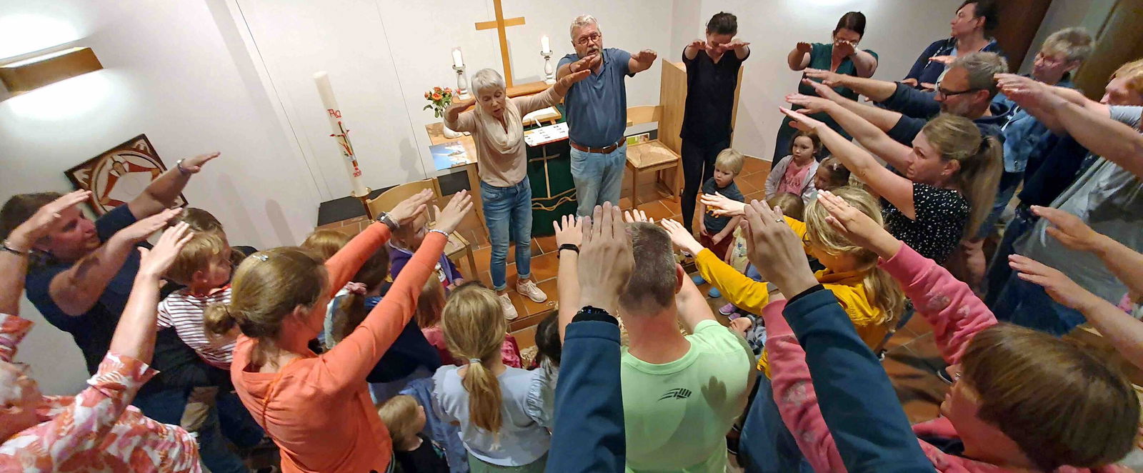 Auf dem Bild feiert eine Gruppe von Menschen einen abendlichen Segen. Sie tragen bunte Sommerkleidung und strecken die Arme nach vorn. Im Hintergrund sieht man ein kleines Holzkreuz auf einem Tisch stehen.
