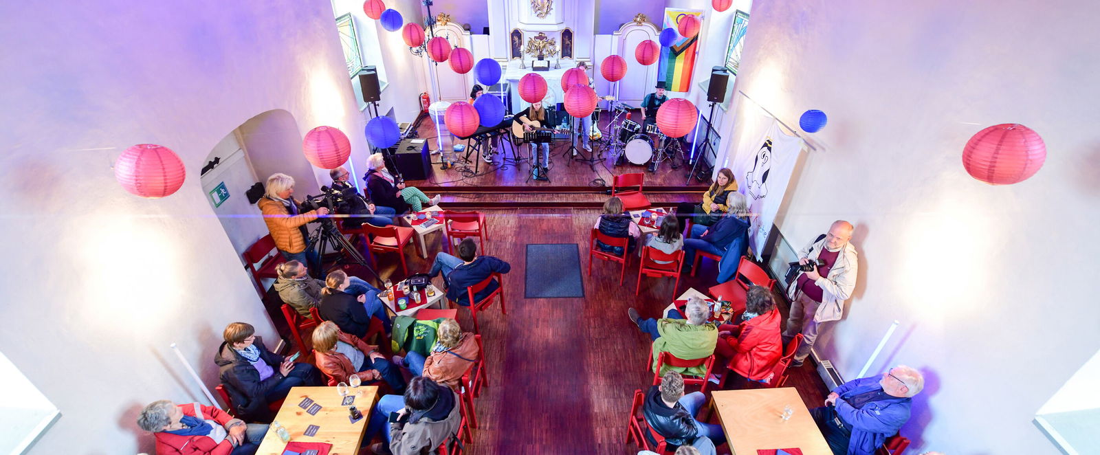 Blick von oben in einen Kirchenraum, in dem bunte Stühle an Tischen stehen, fast alle sind besetzt. Lila, rote und blaue Lampions sind quer durch den Raum gespannt.