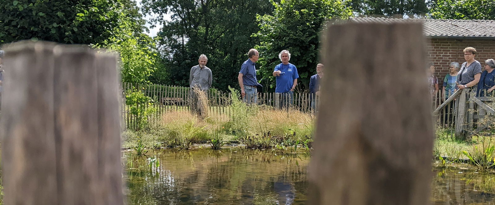 Durch zwei Holzzaun-Pfähle ist ein Teich zu sehen. Am anderen Ufer stehen einige Personen.