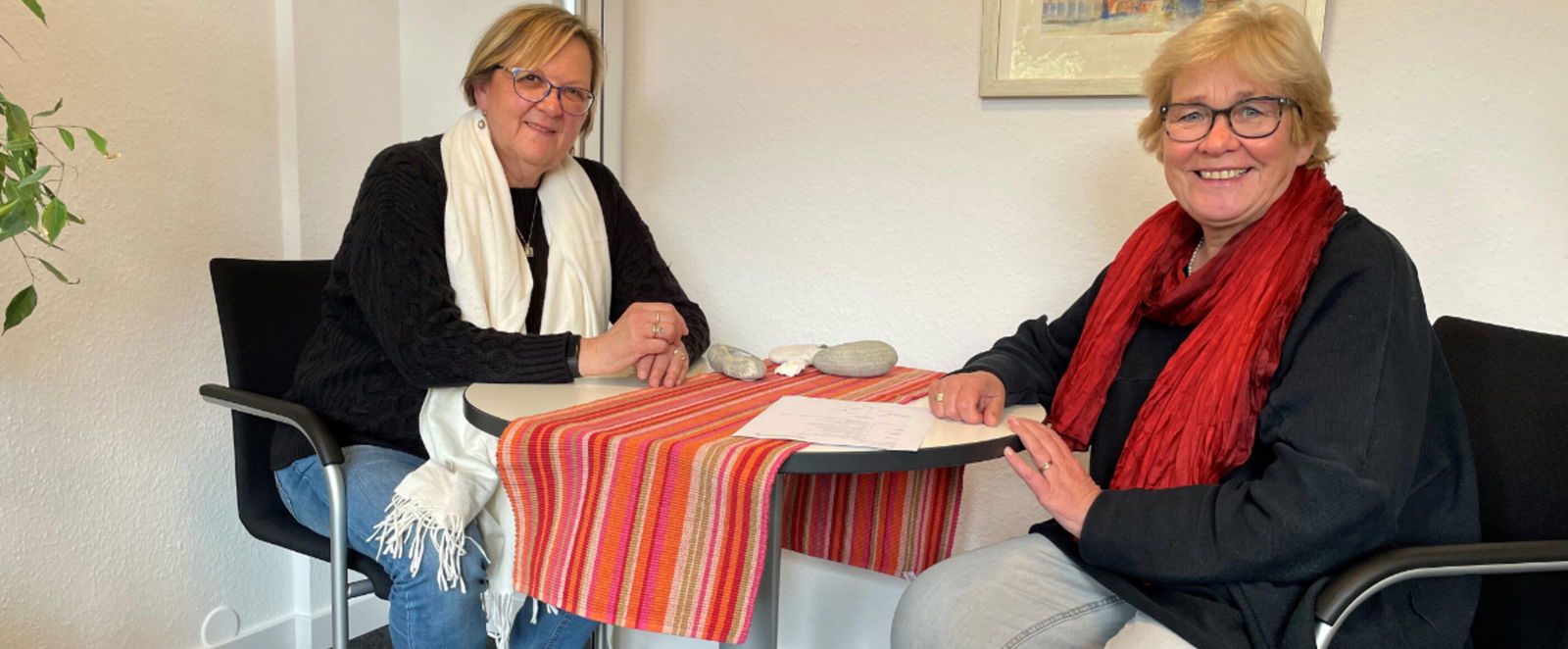 zwei weiblich gelesene Personen sitzen an einem kleinen Besprechungstisch und lächeln freundlich.