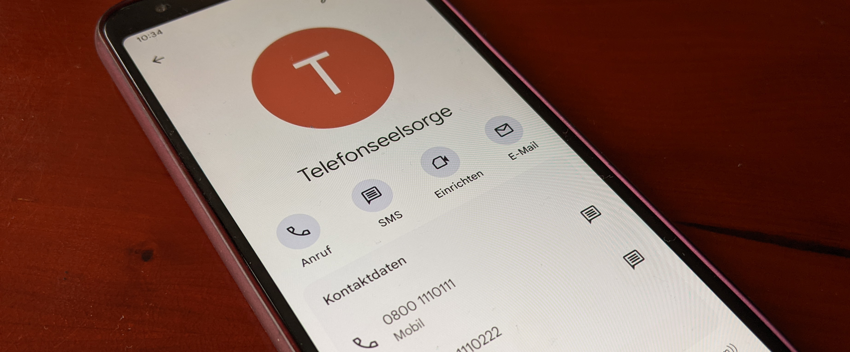 Ein Smartphone liegt auf einem dunklen Holztisch. Es zeigt den Kontakt "Telefonseelsorge" und die Nummern 0800 1110111 und 0800 1110222