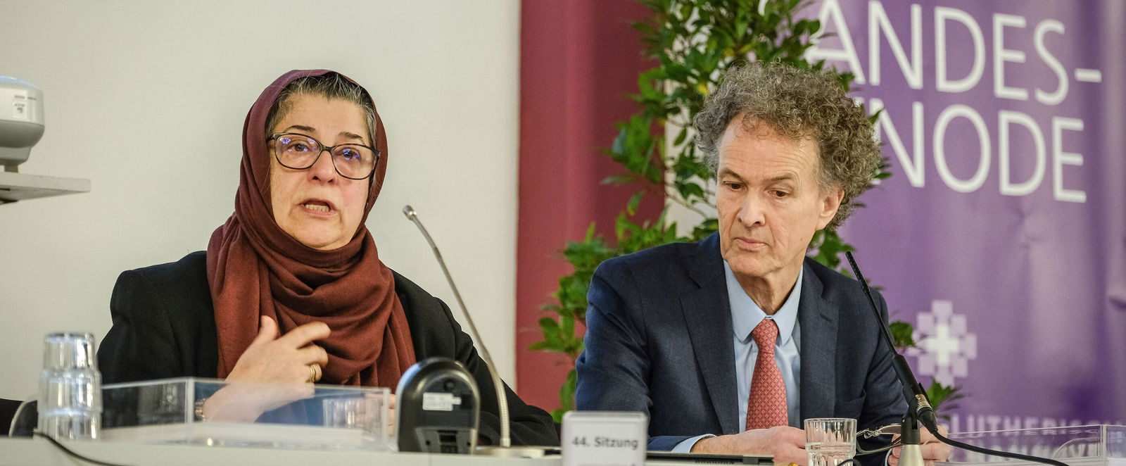 Eine Frau mit Kopftuch und ein Mann im Anzug sitzen an einem Tisch, die Frau gestikuliert. Rechts steht ein Banner mit der Aufschrift "Landessynode"